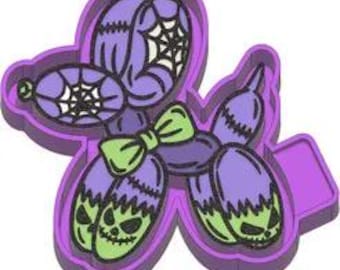 May include: A purple Halloween-themed balloon dog cookie cutter. The design includes a spiderweb, a green bow, and jack-o'-lantern faces. The cutter is made of plastic and has a handle for easy use.