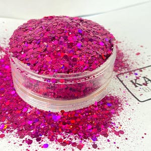 Op de afbeelding: Een doorzichtige plastic container gevuld met levendige magenta glitter. De glitter is een mix van vormen en maten, met wat gemorst op het witte oppervlak. Een label met de tekst "K.A.M. BLANKS" is zichtbaar.