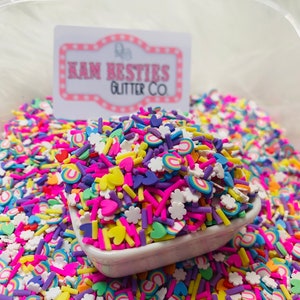 Puede incluir: Un bol blanco lleno de espolvoreados de colores, incluyendo arcoíris, corazones, estrellas y palitos pequeños. Los espolvoreados están rodeados por más espolvoreados en un contenedor más grande. El contenedor tiene una etiqueta que dice "Ram Besties Glitter Co."