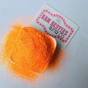 Peut inclure: Gros plan sur un petit bol blanc rempli de paillettes orange vif. Les paillettes sont fines et ont un léger éclat. Le bol est posé sur une surface blanche.