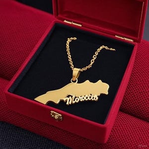 Puede incluir: Un collar dorado con la forma del mapa de Marruecos, presentado en una caja roja. El colgante tiene el nombre del país en escritura cursiva. El collar tiene una cadena dorada y un interior negro.