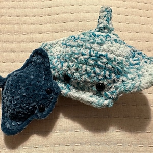 May include: Two plush stingrays, one teal and one dark blue, are displayed. The larger stingray is teal and white, with black button eyes. The smaller stingray is a solid dark blue. Both are made of a soft, textured material.
