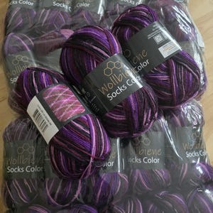 Könnte beinhalten: Mehrere Stränge lila, schwarzer und rosa Sockenwolle werden gezeigt. Das Garn ist mit "Wollbiene Socks Color" beschriftet. Das Garn ist in durchsichtigem Kunststoff verpackt. Das Garn ist eine Mischung aus Farben mit einem Farbverlaufseffekt.