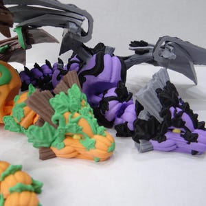 Könnte beinhalten: Eine Sammlung farbenfroher, skurriler Drachenskulpturen. Die Drachen haben kürbisförmige Körper in Orange, Lila und Schwarz, mit grünen Blättern und braunen Flügelstrukturen. Die Flügel sind grau. Diese Fantasiewesen sind perfekt für Halloween.