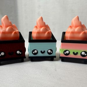 May include: Three colorful, 3D-printed toy dumpsters with flame-shaped toppers. The dumpsters are in maroon, light blue, and striped pink and green. Each has a black rim and base, and a cute face. The flames are orange and peach.