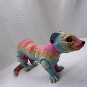 Puede incluir: Un juguete de hurón de colores, pintado con un arcoíris, con extremidades articuladas. El hurón tiene una expresión juguetona y grandes ojos negros.