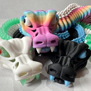 Puede incluir: Una colección de juguetes de serpiente articulados y coloridos. Las serpientes son de varios colores, incluyendo blanco, negro, rosa y verde, con segmentos de arcoíris. Cada serpiente tiene una cabeza detallada con ojos grandes y dientes.