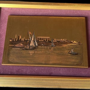 May include: A framed copper relief depicting a river scene with sailboats and buildings, likely Egyptian. The artwork is set against a purple background within a gold-colored frame.