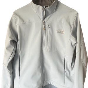 May include: Pale blue Apex jacket with a full-length zip, two side pockets, and the North Face logo on the chest. The jacket is hanging on a green hanger.