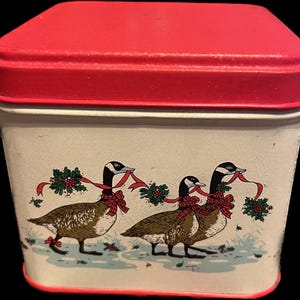 May include: A rectangular tin container with a red lid and trim. The off-white body features a decorative illustration of three geese wearing red bows and holding red ribbons with holly. The container has a vintage, slightly cracked appearance.