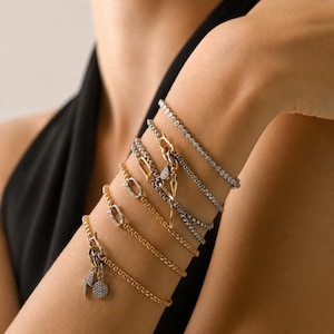 May include: A stack of five bracelets featuring a mix of gold and silver-toned metal chains. One bracelet is a tennis style with clear stones, while others include toggle clasps, a lightning bolt charm, and a circular charm with pavé stones.