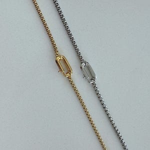 May include: Two fine necklaces, one gold and one silver, are shown against a light backdrop. Each necklace has a delicate chain and a rectangular clasp. The gold necklace is on the left, and the silver necklace is on the right.