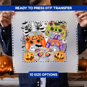May include: A ready to press DTF transfer sheet featuring a Halloween-themed graphic with five video game controllers styled as a skeleton, a green zombie, an orange jack-o'-lantern, a purple witch, and a white mummy, surrounded by ghosts and bats.