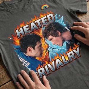 May include: Dark gray t-shirt featuring a graphic of two men facing each other, with the words "HEATED RIVALRY" in red and silver. The design includes flames and ice elements. A rainbow pin is attached to the shirt.