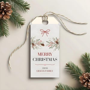 May include: A rectangular Christmas gift tag with a cream background and a decorative bow at the top. The tag features the words "MERRY CHRISTMAS" in burgundy, with "FROM GRACIA FAMILY" below. The tag is tied with a natural twine.