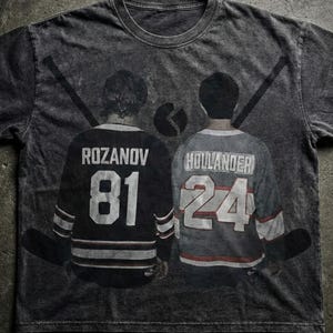 May include: A gray t-shirt featuring a graphic of two hockey players from the back, with their jerseys displaying the names "ROZANOV" and "HOLLANDER" and the numbers "81" and "24". Hockey sticks and a puck are also visible.