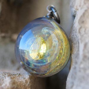 May include: A clear glass pendant with a yellow gemstone center and swirling blue, yellow, and green internal designs. The pendant is attached to a silver-colored metal loop.