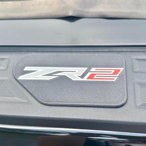 May include: A black rectangular emblem with the letters "ZR2" in white and red. The emblem is set against a textured black surface, likely part of a vehicle's exterior.
