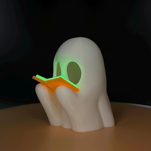 May include: A white ghost-shaped figurine with a glowing green interior and a small orange book. The ghost is reading the book, which is held in its hands. The background is dark.