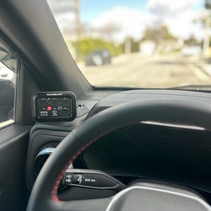 May include: Black control panel labeled "AUXBEAM" mounted on a car's dashboard. The panel has illuminated icons for various functions, including lights. The steering wheel is visible in the foreground, with a red stitched detail.