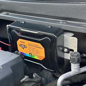 May include: A black RGB multifunction switch assembly, model RA60, mounted in an engine compartment. The assembly features a silver mounting bracket and a label with technical specifications, including voltage and wattage. The label also includes the brand name AUXBEAM.