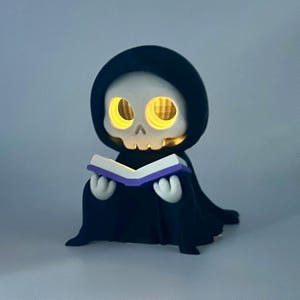May include: A novelty light-up figurine of a cartoon Grim Reaper reading a book. The figure is black with a white skull face and glowing yellow eyes. The book has a purple cover. A unique decorative item for Halloween.