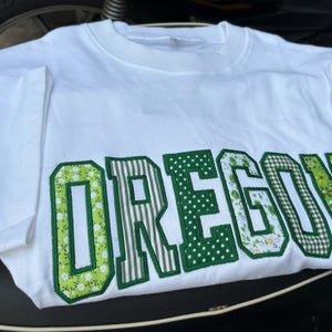 May include: A white short-sleeved t-shirt featuring the word OREGON in large block letters across the chest. Each letter is filled with a different green patterned fabric, including floral, striped, polka dot, and gingham prints, outlined in green.