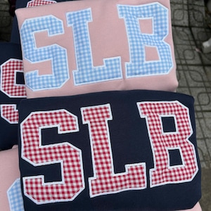 May include: A stack of folded sweatshirts featuring large block letters that spell SLB. The top sweatshirt is light pink with light blue gingham patterned letters. The middle sweatshirt is navy blue with red gingham patterned letters.