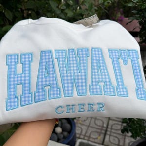 May include: A white crewneck sweatshirt featuring the word HAWAI'I in large, light blue and white gingham-patterned block letters with a light blue embroidered outline, and the word CHEER embroidered in light blue letters underneath.