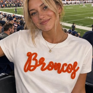 May include: A white short-sleeved t-shirt featuring the word Broncos embroidered in orange yarn across the chest. The shirt is paired with light blue denim jeans. The design includes a crew neckline and a standard fit, showcasing a textured script.