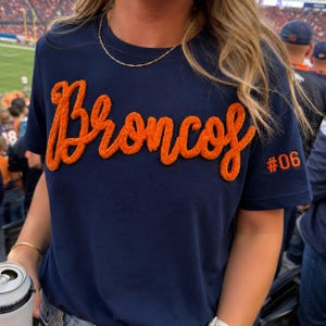 May include: A navy blue short-sleeved t-shirt featuring the word Broncos in large, orange, textured chenille-style lettering across the chest. The left sleeve includes the orange embroidered text #06. The shirt has a crew neckline.