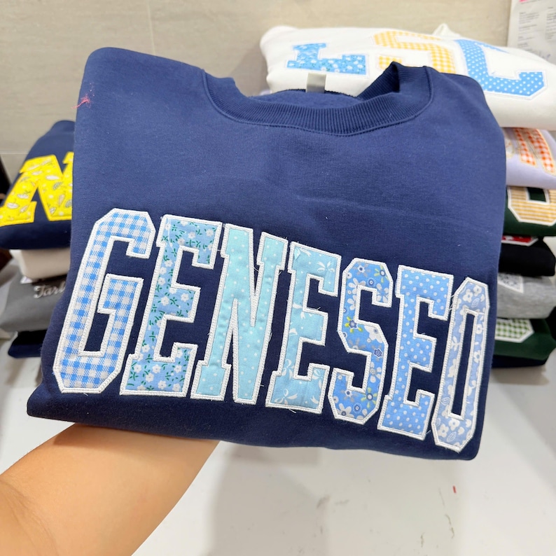 May include: A navy blue sweatshirt with the word "GENESEO" appliqued across the front in a patchwork style. The letters feature various light blue and white patterns, including gingham and floral prints. Additional sweatshirts are stacked in the background.