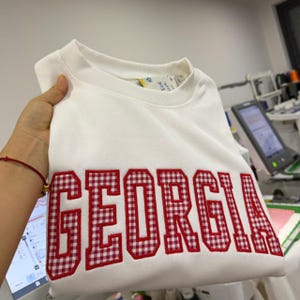 May include: White sweatshirt with the word "GEORGIA" in large, red and white gingham letters across the front. The sweatshirt has a crew neck and long sleeves. The garment is folded and held by a hand.