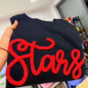 May include: A navy blue crewneck sweatshirt featuring the word Stars in a large, raised, red textured script font across the front. The lettering has a fuzzy, yarn-like appearance that stands out against the smooth dark fabric of the garment.