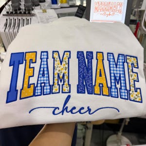 May include: A white sweatshirt featuring the embroidered text TEAM NAME in a collegiate font with alternating blue, yellow, and gingham patterns. Below the main text, the word cheer is embroidered in a blue cursive script font.