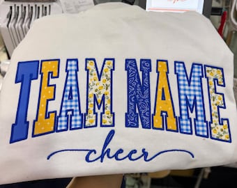 May include: A white sweatshirt featuring the embroidered text TEAM NAME in a collegiate font with alternating blue, yellow, and gingham patterns. Below the main text, the word cheer is embroidered in a blue cursive script font.