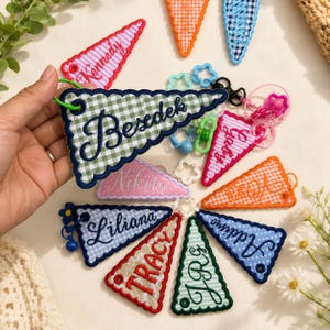 May include: A collection of triangular fabric pennant keychains featuring gingham patterns in various colors. Each pennant is embroidered with a name in cursive script, including Besdek, Kennedy, Gaby, Nikita, Liliana, Tracy, and Addie, with plastic rings.