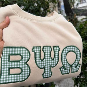 May include: A beige crewneck sweatshirt featuring large, embroidered Greek letters that read BΨΩ. The letters are filled with a green and white gingham check pattern and are outlined with a thick, dark green stitched border.