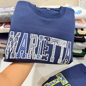 May include: A navy blue sweatshirt with the word "MARIETTA" across the front in a patchwork design. The letters feature various patterns, including gingham, floral, and polka dots. Other sweatshirts are stacked in the background.