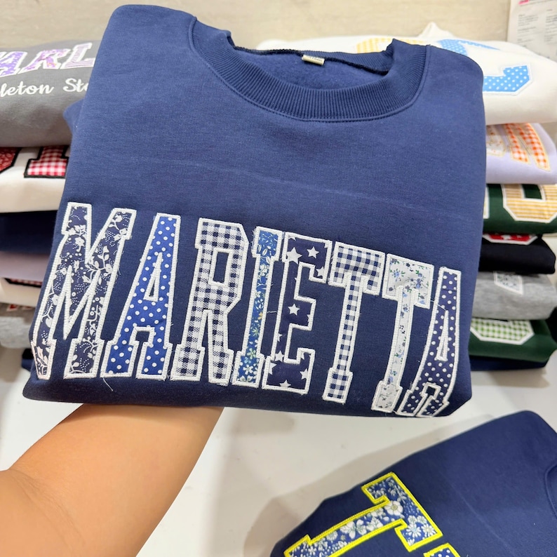 May include: A navy blue sweatshirt with the word "MARIETTA" across the front in a patchwork design. The letters feature various patterns, including gingham, floral, and polka dots. Other sweatshirts are stacked in the background.