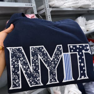 May include: Navy blue sweatshirt with the letters "NYMT" in white. The "N" and "Y" are filled with a floral pattern, the "M" has blue and white stripes, and the "T" has white stars. The sweatshirt is folded.