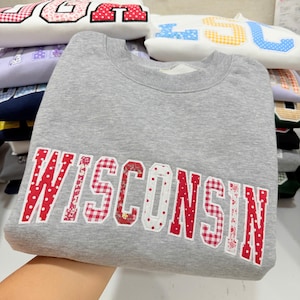 May include: A gray sweatshirt with the word "WISCONSIN" appliqued in red fabric with white polka dots, floral, and gingham patterns. Other sweatshirts with different letters are stacked in the background.