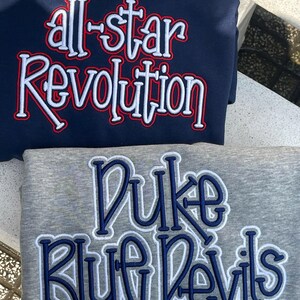 May include: Two sweatshirts, one navy blue and one gray. The navy blue sweatshirt has the words "all-star Revolution" embroidered in white with red accents. The gray sweatshirt has the words "Duke Blue Devils" embroidered in blue and white.