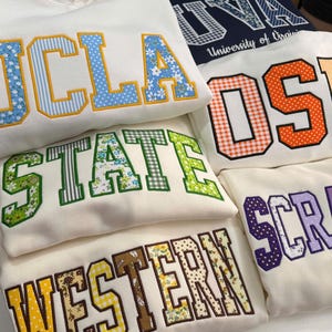 May include: A collection of white sweatshirts featuring large, block-letter collegiate-style appliqués. The letters are filled with various colorful patterns, including floral prints, gingham, and polka dots, outlined in bold, contrasting thread.