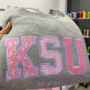 May include: A gray sweatshirt with the letters "KSU" appliqued in pink and white patterned fabric, outlined in purple stitching. The word "mom" is embroidered below the letters. The sweatshirt is folded.