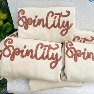 May include: A stack of folded cream-colored sweatshirts featuring the text SpinCity embroidered in a raised, textured, terracotta-colored yarn. The cursive lettering is applied across the front of each garment in a consistent style.