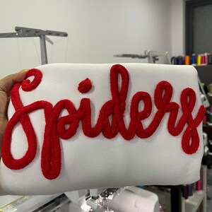 May include: A white sweatshirt featuring the word Spiders embroidered in a large, bold, red cursive font. The lettering has a thick, textured, fuzzy appearance that stands out against the smooth, plain white fabric of the garment.