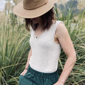 Könnte beinhalten: Eine Frau trägt ein weißes Stricktop mit V-Ausschnitt und einen grünen Rock mit Knöpfen. Sie trägt einen braunen Hut und steht in einem Feld mit hohem Gras.