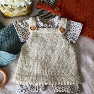 May include: A white knitted baby overall with two wooden buttons. The overall is worn over a white and blue floral print short-sleeved shirt.