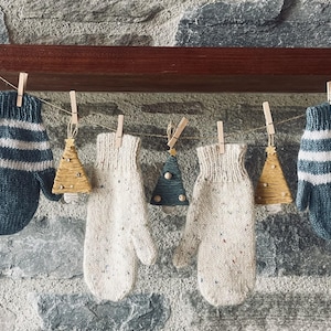 May include: A display of handmade mittens and Christmas tree ornaments hanging from a string. The mittens are blue with white stripes and speckled off-white. The ornaments are small, wooden Christmas trees in gold and teal.